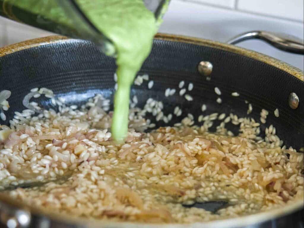 A close-up of a pan with sautéed onions and uncooked rice, while a green liquid is being poured in from a blender, likely for a risotto recipe.